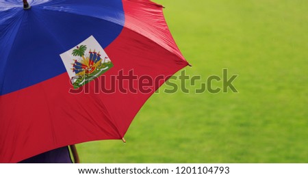 Haiti flag umbrella. Closeup of printed umbrella over green grass field background. Landscape, side view. Rainy weather/ climate change and global warming concept.