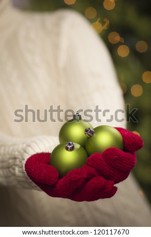 Woman Wearing A Sweater and Seasonal Red Mittens Holding Three Green Christmas Ornaments.