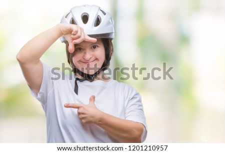 Young adult cyclist woman with down syndrome wearing safety helmet over isolated background smiling making frame with hands and fingers with happy face. Creativity and photography concept.