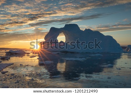Sunburst behind a massive iceberg with a hole in it during the midnight sun season. Disko bay, Greenland. Royalty-Free Stock Photo #1201212490