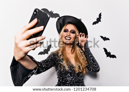 Beautiful curly witch making selfie in halloween. Joyful caucasian girl taking picture of herself at vampire party.