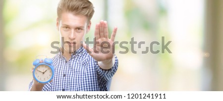 Young handsome blond man holding alarm clock with open hand doing stop sign with serious and confident expression, defense gesture