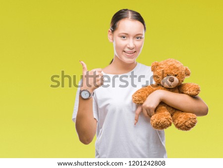 Young caucasian woman holding teddy bear over isolated background happy with big smile doing ok sign, thumb up with fingers, excellent sign