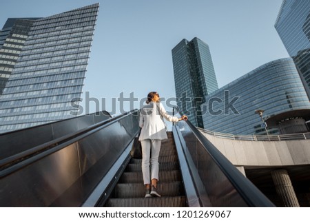 Stylish businesswoman in white suit going up on the escalator at the business centre outdoors with skyscrapers on the background in Paris Royalty-Free Stock Photo #1201269067
