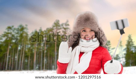 people, technology and leisure concept - happy smiling woman in fur hat taking picture by smartphone on selfie stick over winter forest background