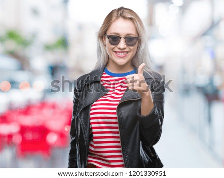 Young blonde woman wearing fashion jacket and sunglasses over isolated background doing happy thumbs up gesture with hand. Approving expression looking at the camera with showing success.