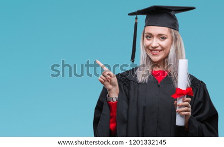 Young blonde woman wearing graduate uniform holding degree over isolated background very happy pointing with hand and finger to the side
