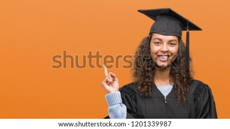 Young hispanic woman wearing graduation uniform very happy pointing with hand and finger to the side