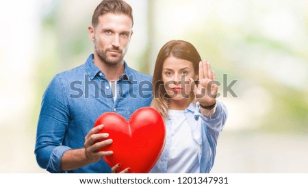 Young couple in love holding red heart over isolated background with open hand doing stop sign with serious and confident expression, defense gesture