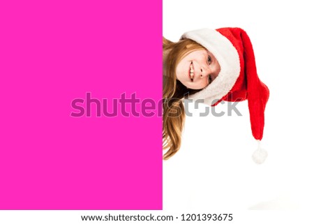 Little red haired and freckled face girl dressed in Santa's hat holds blank flipchart board and dreams about gifts. Christmas and New Year advertising concept. Isolated on a blurred white background