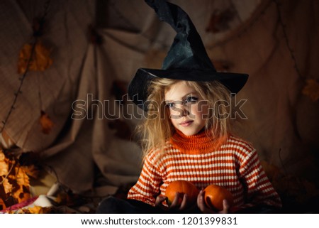 little happy cute shaggy with tangled hair witch 5 -6 years old laughing in black sorcerer hat portrait on dark bright background with pumpkin, halloween, sinister environment