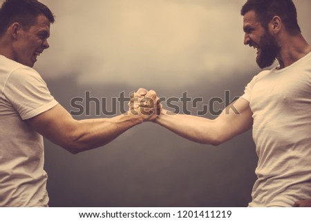 Two crossfit sports men doing arm wrestling exercise during outdoor workout against cloudy sky and hills background.