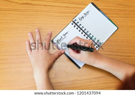 Woman fills in her goals and plans for long and short terms in spiral notepad on wooden table, top view.