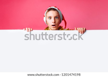 Portrait of a beautiful teen in Santa's hat and with tinsel on her neck, posing behind a white panel isolated on a red background. Teen baby holding an empty Billboard. Christmas