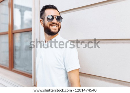 Stylish hipster in a white t-shirt with sunglasses and a beard, standing near a white wooden wall