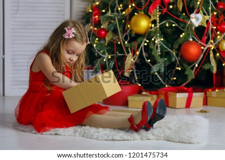 Little girl in red dress sits on carpet and opens a gift. Christmas tree with ornaments in the background. Happy childhood.
