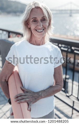 Waiting for you. Pretty elderly female embracing yoga mat and looking forward while keeping smile on her face
