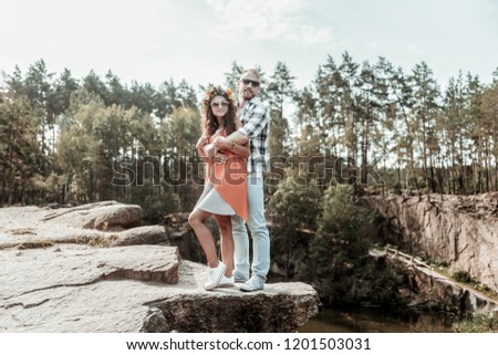 Near beloved. Beautiful stylish curly woman wearing sunglasses standing near her beloved while standing on the rock near lake
