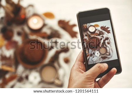 Hand holding phone and taking photo of stylish autumn flat lay of coffee,cookies, fall leaves, candle, berries, nuts, acorns,cotton on soft white background. Modern food photography