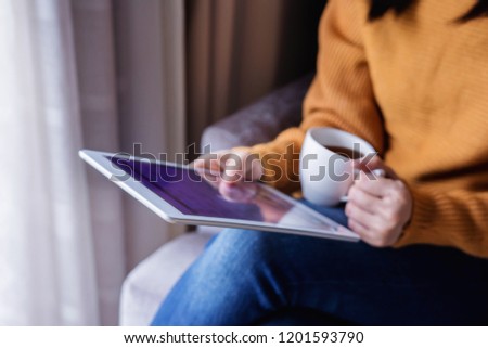Reading Concept. Young Woman using Tablet and Drinking Coffee. Relaxing in Cozy House, Sit on the Chair, Front View and Selective Focus