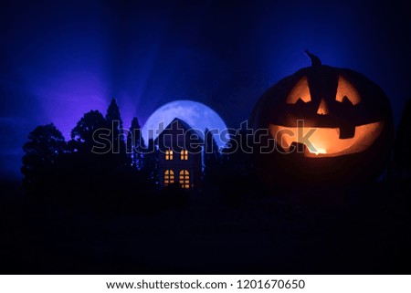 Horror view of Halloween pumpkin with scary smiling face. Head jack lantern with haunted building and tree on dark toned foggy background.