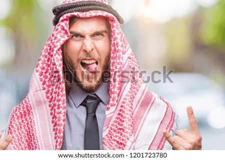 Young handsome arabian man with long hair wearing keffiyeh over isolated background shouting with crazy expression doing rock symbol with hands up. Music star. Heavy concept.