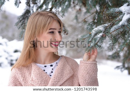 Happy blonde girl in a pink hat and sweater with gloves in the winter forest is smiling. The girl looks at the branch of spruce. Beauty photo. Close-up. Portrayed orientation.