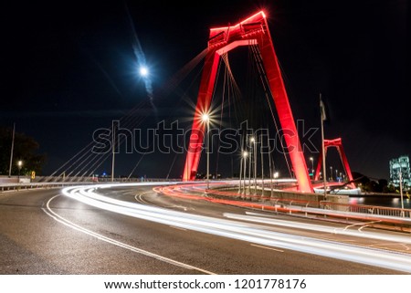 Lighttrails on a road bend in front of a red bridge named Willemsbrug in Rotterdam at night with a full moon.