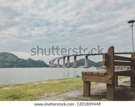 A empty bench with a bridge and the water In the background
