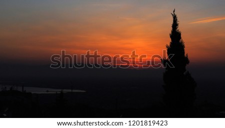 A sunset view across the city and across the sea to Mt Olympus from the Castle area in Thessaloniki, Greece, in fiery red and orange colors, with a silhouette of a tree in the foreground.