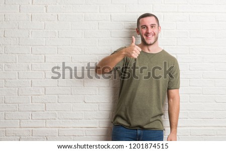 Young caucasian man standing over white brick wall doing happy thumbs up gesture with hand. Approving expression looking at the camera with showing success.