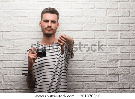 Young man holding vintage camera standing over white brick wall with angry face, negative sign showing dislike with thumbs down, rejection concept