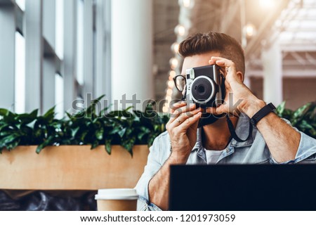 Young bearded hipster man with glasses and denim jacket sits in coffee shop at table in front of laptop and takes instant photo on camera. Tourist takes picture from the window.