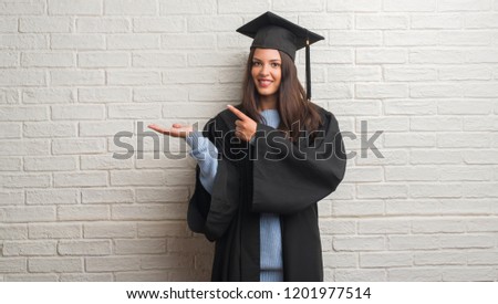 Young brunette woman standing over white brick wall wearing graduate uniform very happy pointing with hand and finger