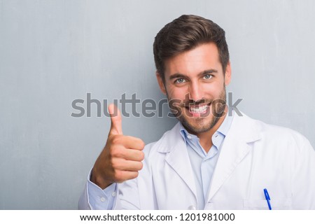 Handsome young professional man over grey grunge wall wearing white coat doing happy thumbs up gesture with hand. Approving expression looking at the camera with showing success.