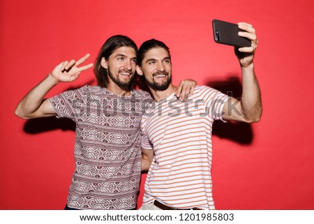 Portrait of a two young happy twin brothers isolated over red background, using mobile phones, taking a selfie