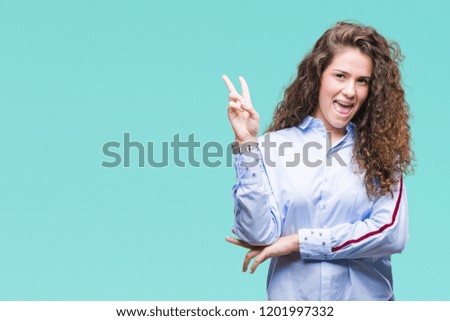 Beautiful brunette curly hair young girl wearing elgant look over isolated background smiling with happy face winking at the camera doing victory sign. Number two.