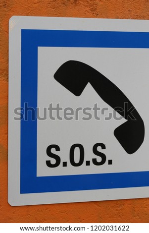 Close up view of a french telephone symbol with the sign SOS to prevent danger. Black sign with a blue frame and white background. Emergency pictogram fixed on a orange painted wall.