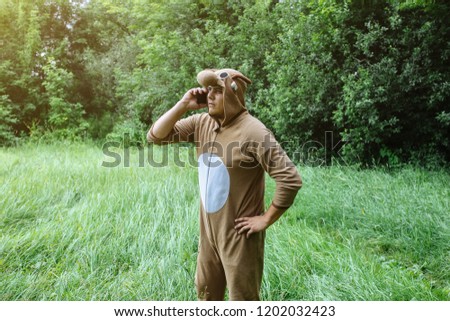 Young man is standing in the forest in cosplay costume of a cow. Guy in the funny animal pyjamas sleepwear is talking on smartphone outdoors. Halloween ideas for party in the nature.