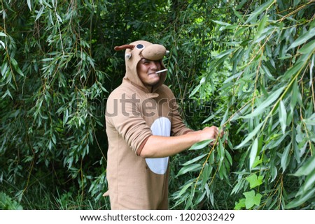 Young man is smoking in the forest in cosplay costume of a cow. Guy in the funny animal pyjamas sleepwear in the nature. Halloween ideas for party.