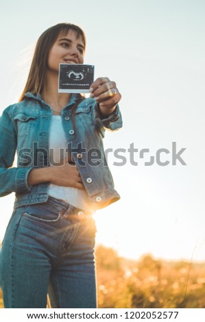 Young Pregnant Woman holding ultrasound photo at Sunset and Embracing her Belly. 4 Month Pregnancy. Maternity Concept. Toned Photo.