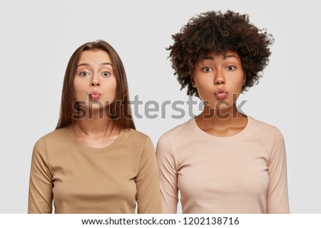 Horizontal shot of multiracial girls make kiss with lips, flirt with handsome guys, express tenderness and calmness, dressed in casual clothes, isolated over white studio wall. Multiethnic women