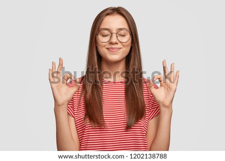 Pleased satisfied young female model makes zero gesture, wears transparent glasses, has long dark hair, dressed in casual striped t shirt, stands over white background. People, body language concept Royalty-Free Stock Photo #1202138788