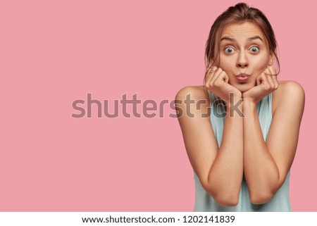 Horizontal shot of beautiful dark haired woman keeps hands under chin and purses lips for making kiss, has stunned expression and bugged eyes, poses over pink background with free space aside on left