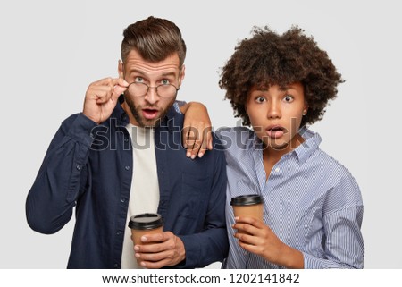 Picture of astonished mixed race woman and man stare with surprised epressions at camera, cannot believe own eyes, drink coffee, pose against white background together. Interracial couple indoor
