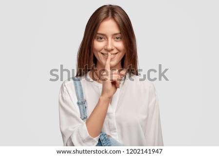 Shh, its privacy! Good looking dark haired woman with charming smile, keeps index finger over mouth, shows hush gesture, gossips with best friend about boyfriends, dressed in fashionable clothes Royalty-Free Stock Photo #1202141947
