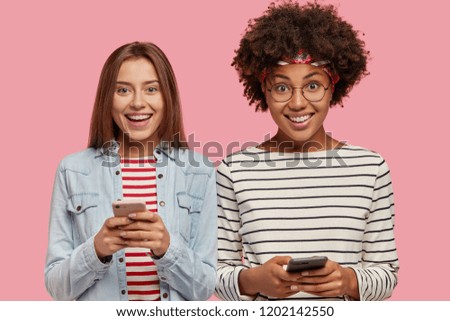 Horizontal view of happy interracial addicted teenagers hold modern cellulars in hands, play online games or chat in social networks, isolated over pink background, read pleasant notifications