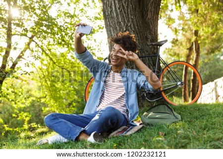 Cheerful young african teenager with backpack outdoors, sitting on grass, taking a selfie with mobile phone