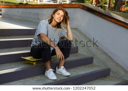Photo of happy skater guy 16-18 in casual wear sitting on skateboard in skate park during sunny summer day