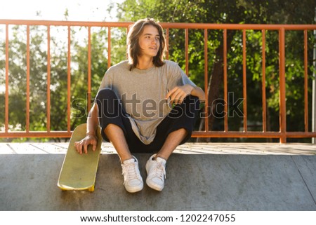 Photo of european skater boy 16-18 in casual wear sitting on ramp with skateboard in skate park during sunny summer day